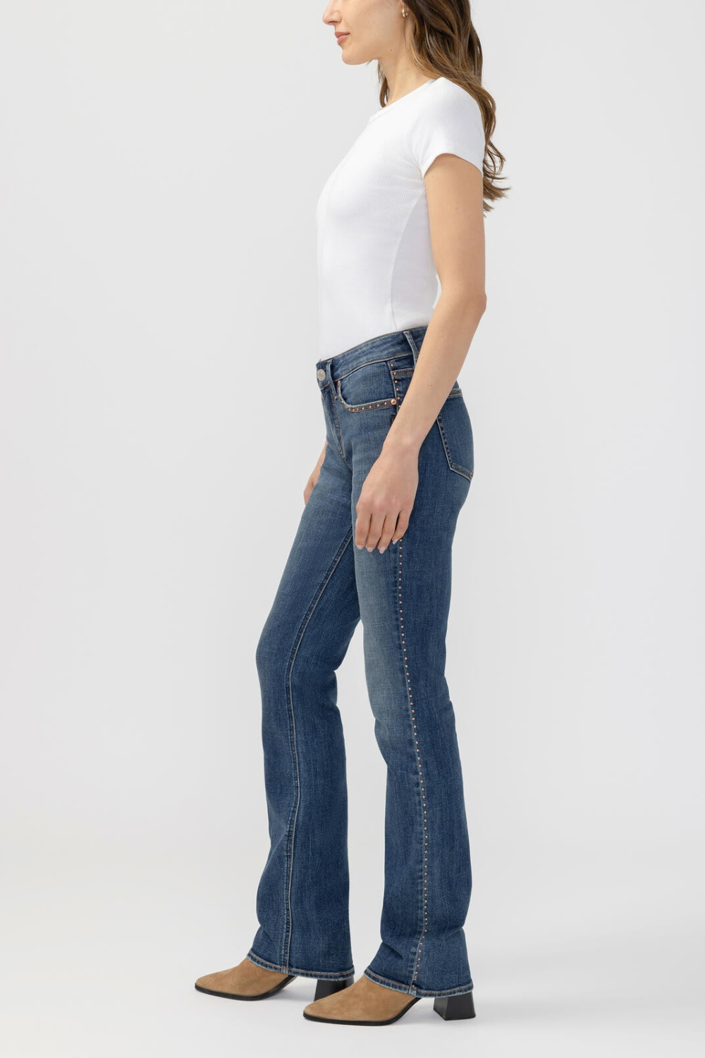 A woman shown in side profile wearing blue denim jeans with decorative studs down the outer seam, a white short-sleeve top, and brown heeled boots against a light gray studio background.