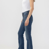 A woman shown in side profile wearing blue denim jeans with decorative studs down the outer seam, a white short-sleeve top, and brown heeled boots against a light gray studio background.