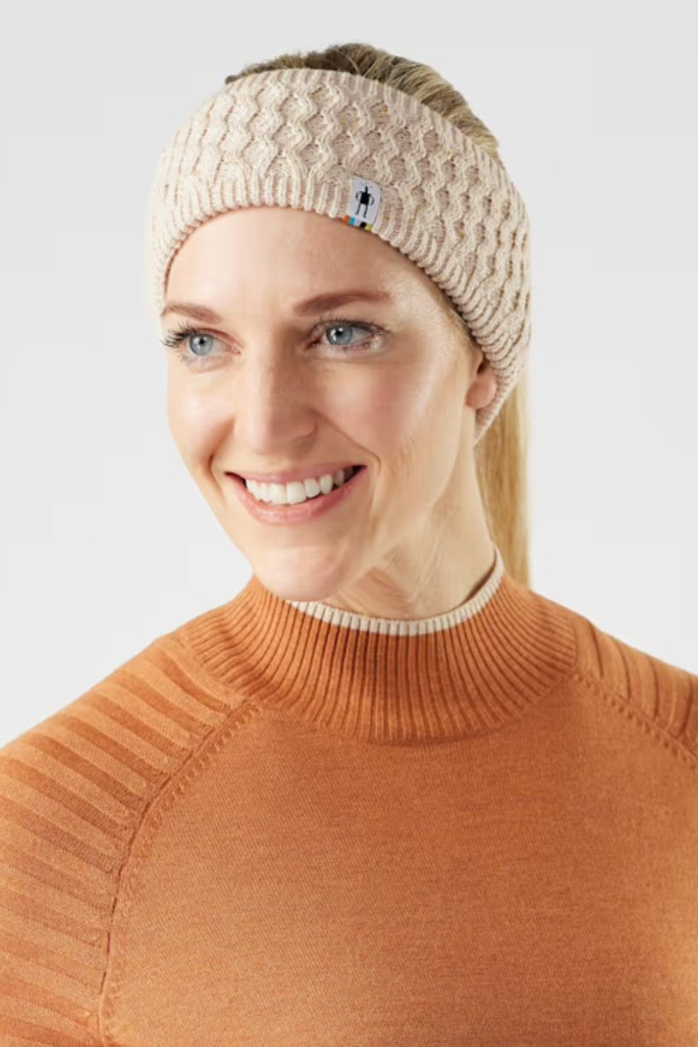 Woman wearing a beige knitted headband with a brand logo on a light gray background