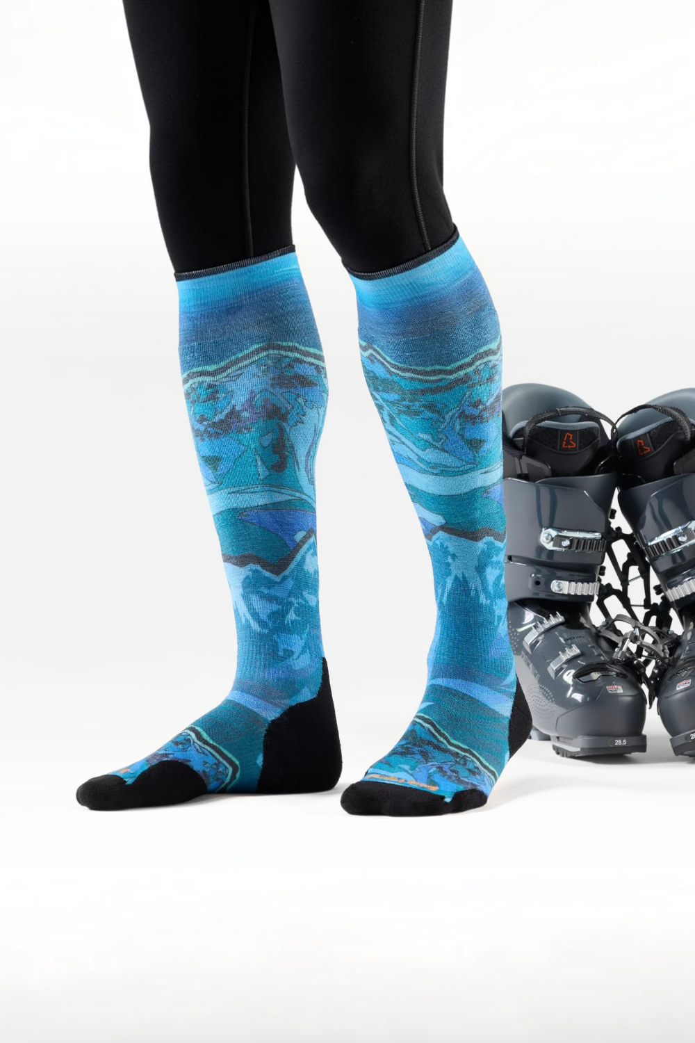 A person wearing blue and turquoise abstract-patterned ski socks with black toe and heel sections, paired with black leggings. A pair of black ski boots sits on a white studio background.