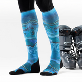 A person wearing blue and turquoise abstract-patterned ski socks with black toe and heel sections, paired with black leggings. A pair of black ski boots sits on a white studio background.