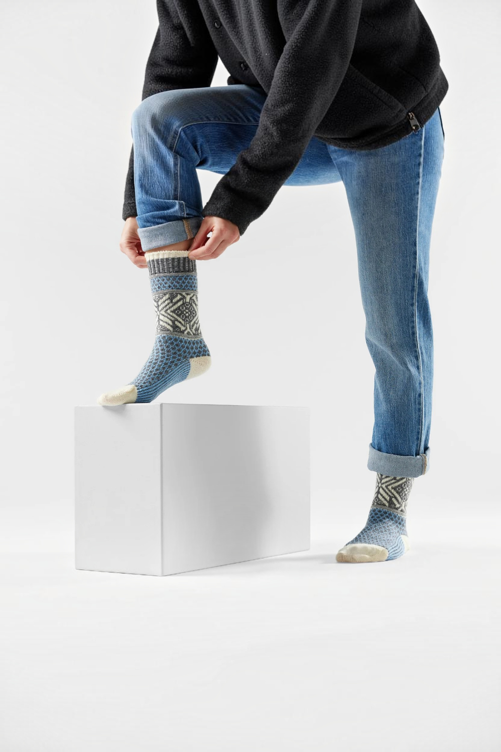 A person wearing blue jeans and a dark cardigan sits on a white block, adjusting a pair of patterned blue knit socks with cream toes and heels against a clean white studio background.