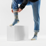 A person wearing blue jeans and a dark cardigan sits on a white block, adjusting a pair of patterned blue knit socks with cream toes and heels against a clean white studio background.