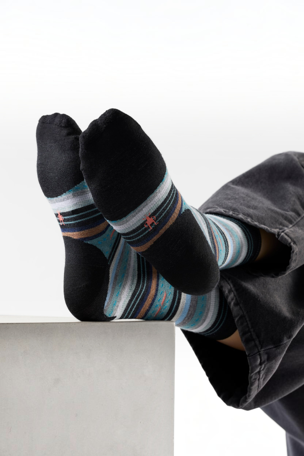 A person's feet wearing black crew socks with blue, gray, and brown striped patterns and small red star accents, resting on a light gray block against a white background.