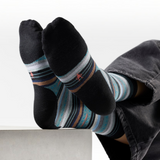 A person's feet wearing black crew socks with blue, gray, and brown striped patterns and small red star accents, resting on a light gray block against a white background.