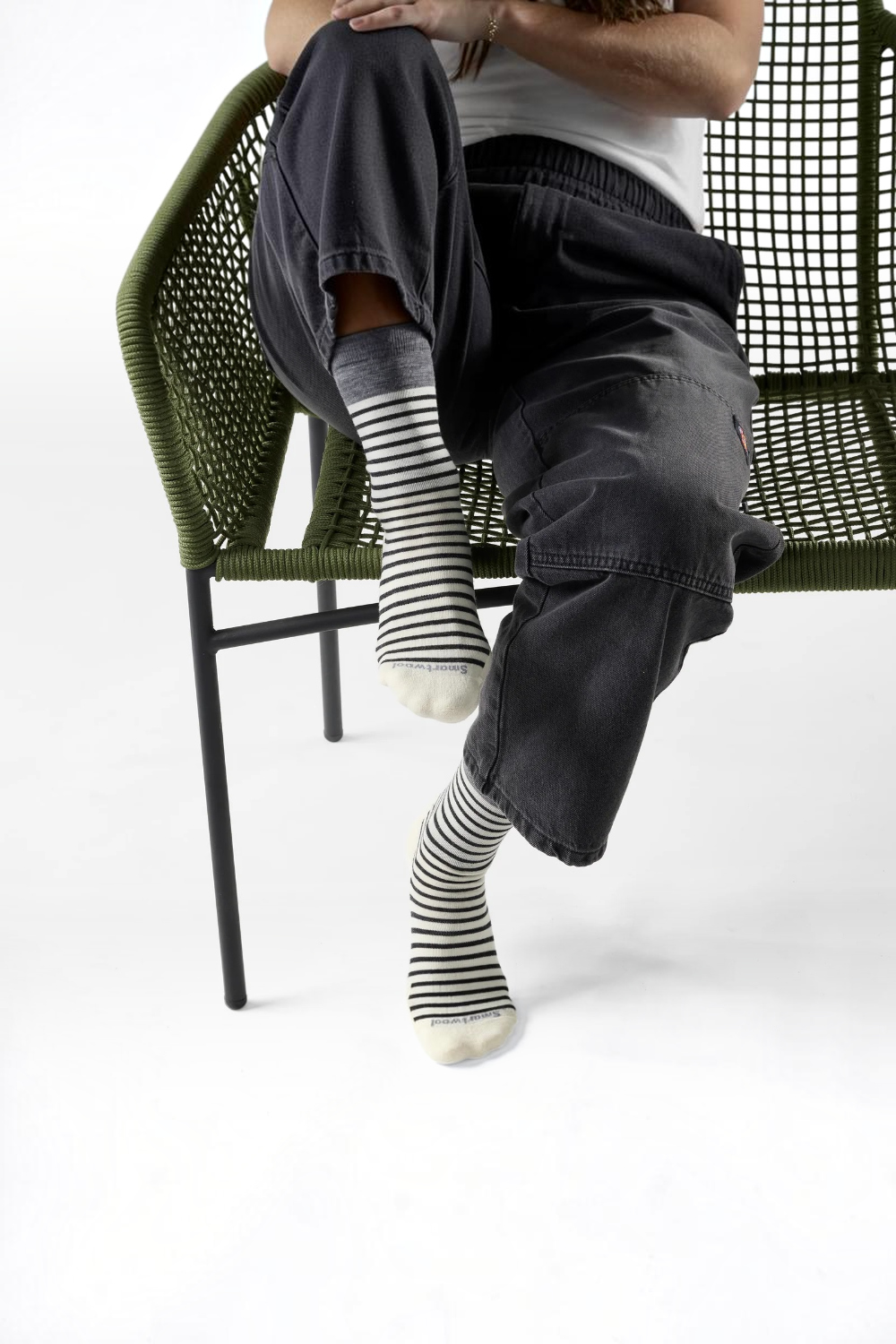 A person seated on a green woven chair wearing black-and-white striped crew socks, dark charcoal pants, and a white top against a clean white studio background.