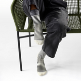 A person seated on a green woven chair wearing black-and-white striped crew socks, dark charcoal pants, and a white top against a clean white studio background.