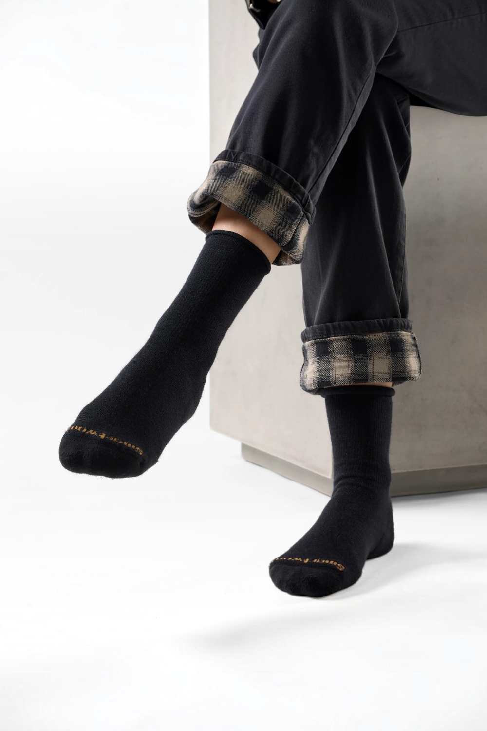 A person seated on a light gray bench wearing black crew socks with small gold text near the toes and rolled black-and-gray plaid pants, photographed in a clean studio setting.