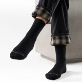 A person seated on a light gray bench wearing black crew socks with small gold text near the toes and rolled black-and-gray plaid pants, photographed in a clean studio setting.