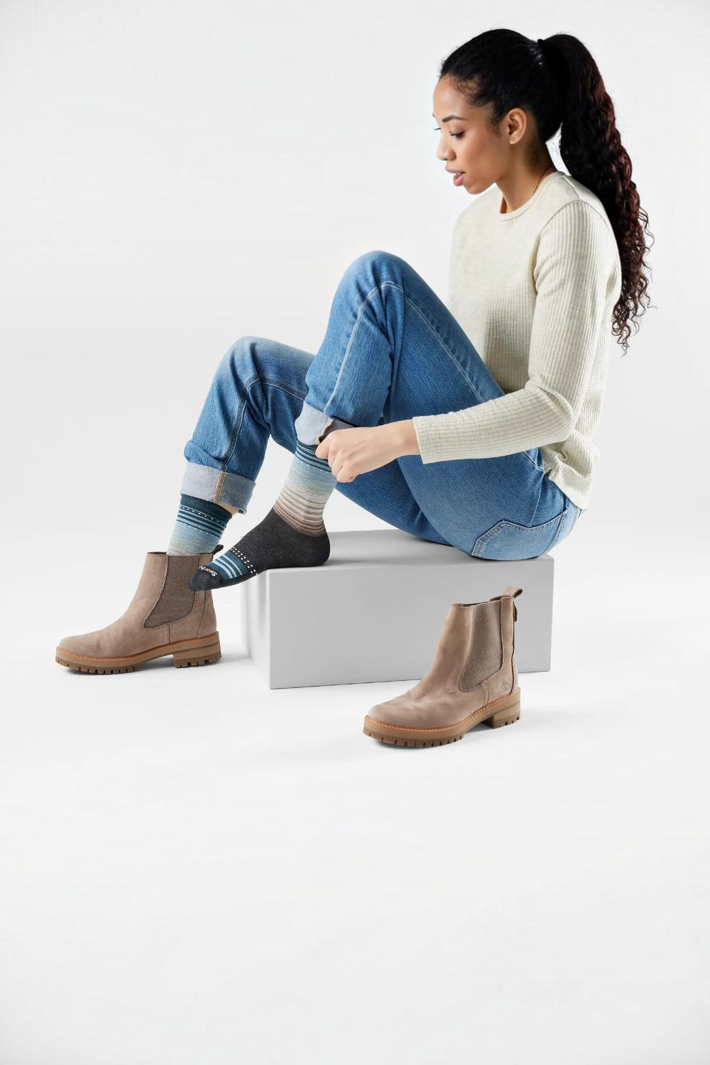 Woman wearing a long sleeve white top and blue jeans sitting on a box putting on a crew length sock with a mostly black foot and tan, grey and blue stripes on a light background.