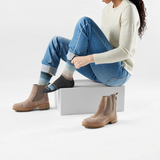 Woman wearing a long sleeve white top and blue jeans sitting on a box putting on a crew length sock with a mostly black foot and tan, grey and blue stripes on a light background.