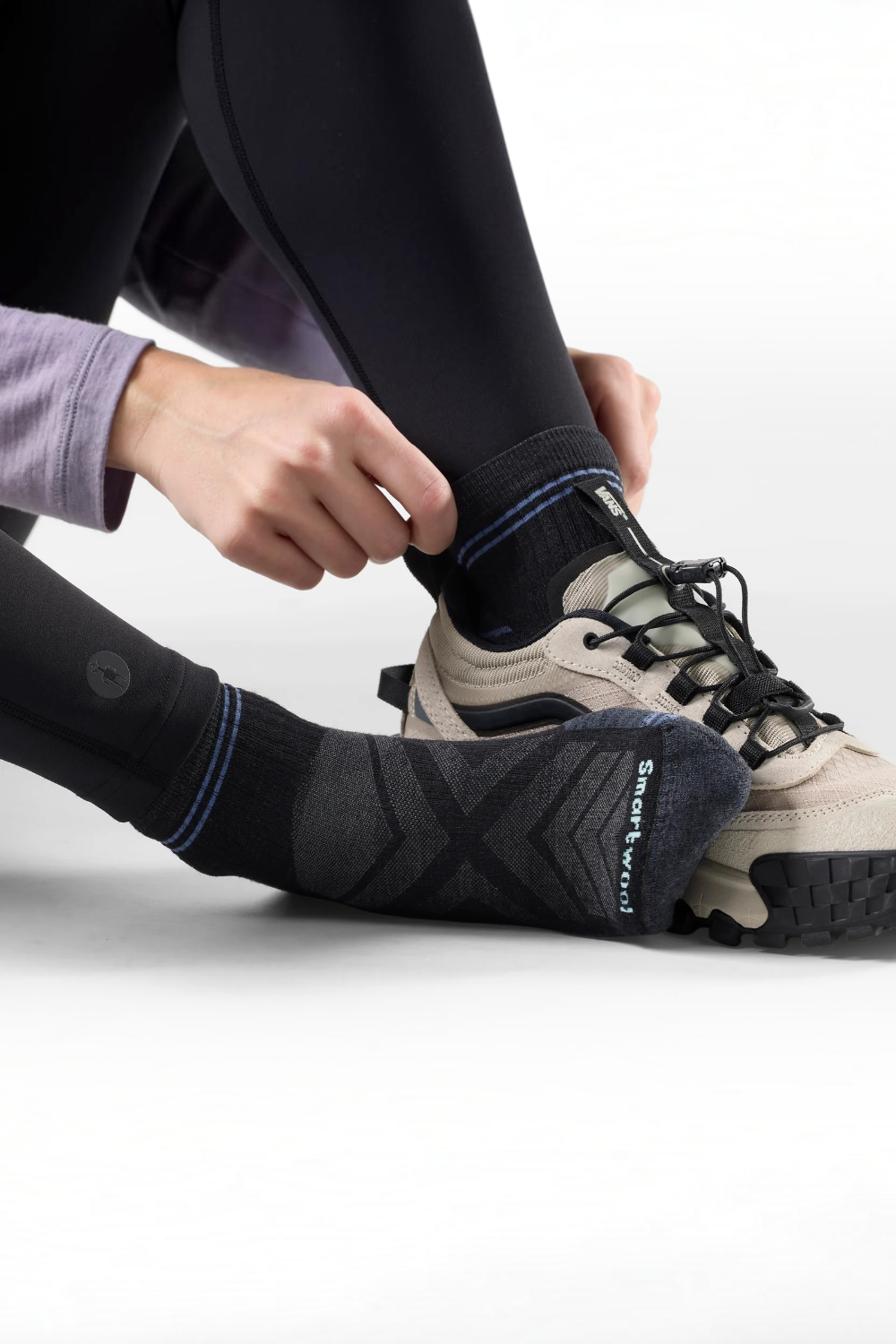 A close-up of a person wearing black athletic socks with purple stripe accents and lace-up sneakers, adjusting the front sock. The socks have a textured knit design and the brand name 'Smartwool' is visible near the toe.