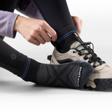 A close-up of a person wearing black athletic socks with purple stripe accents and lace-up sneakers, adjusting the front sock. The socks have a textured knit design and the brand name 'Smartwool' is visible near the toe.