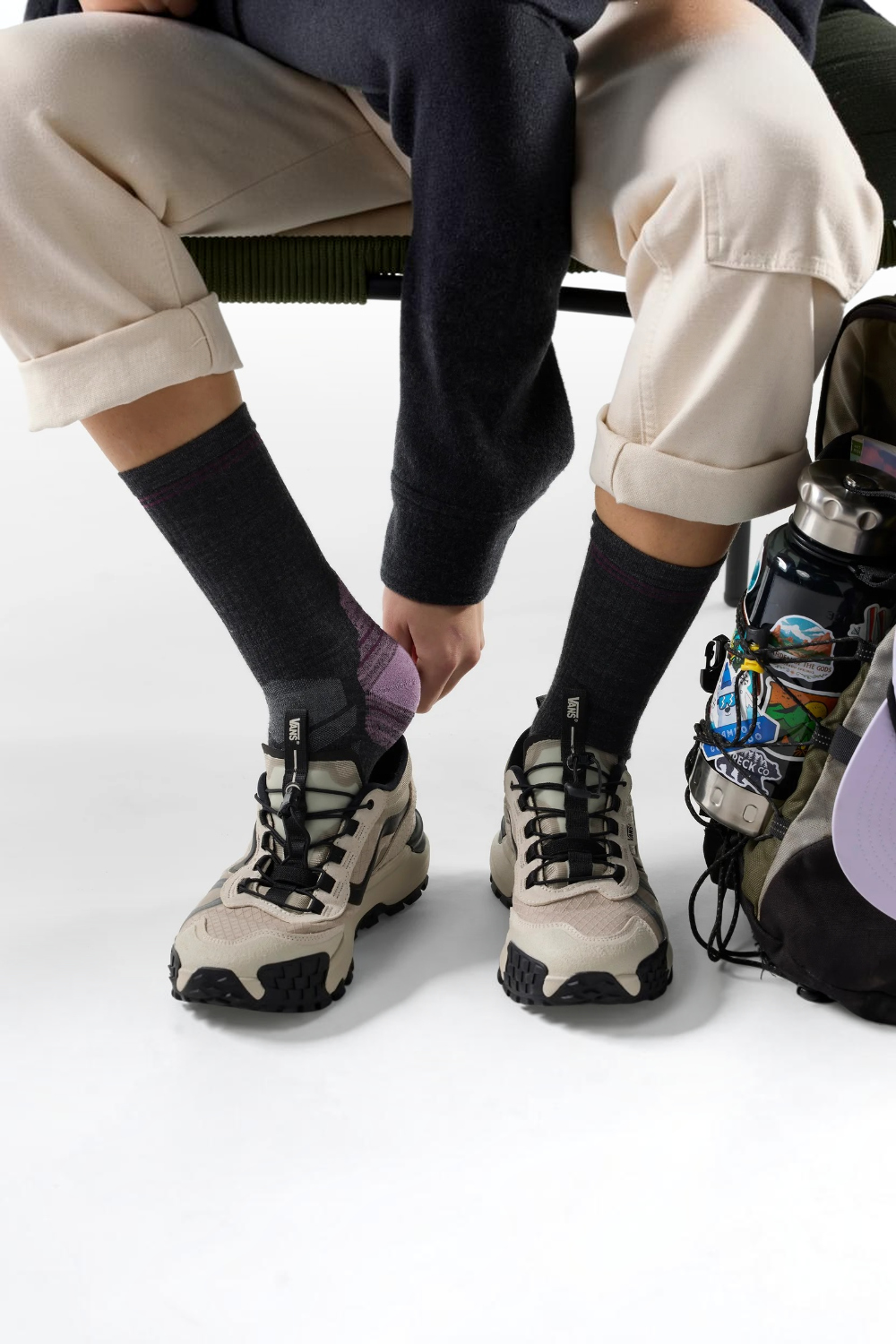 A person seated on a bench wearing beige cargo pants, dark socks, and chunky beige-and-black hiking shoes, with a water bottle bag beside them.