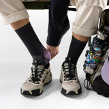A person seated on a bench wearing beige cargo pants, dark socks, and chunky beige-and-black hiking shoes, with a water bottle bag beside them.
