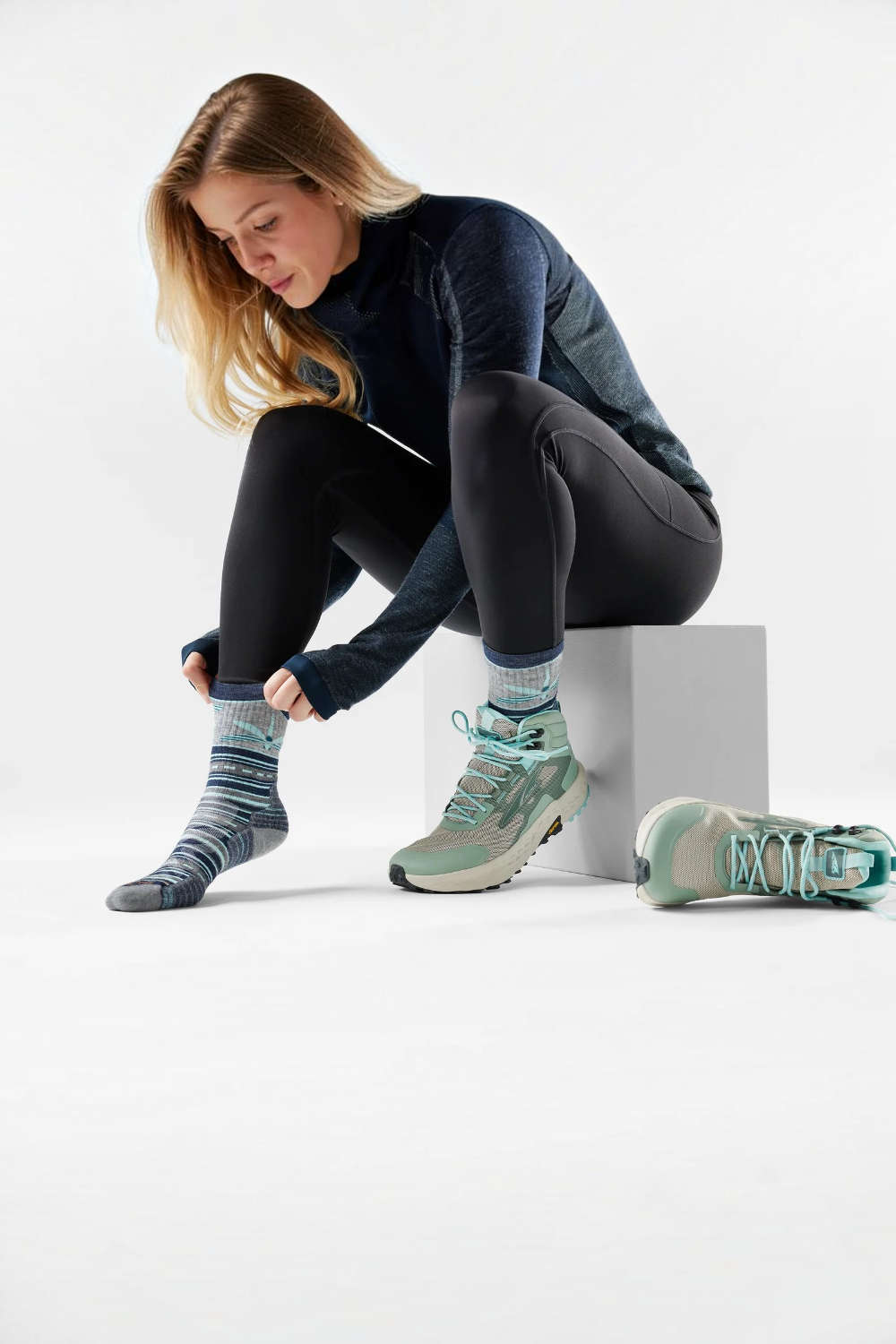 A woman sits on a white block in a studio setting, putting on striped athletic socks. She is wearing a dark blue long-sleeve top, black leggings, and mint green trail running shoes.