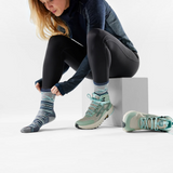 A woman sits on a white block in a studio setting, putting on striped athletic socks. She is wearing a dark blue long-sleeve top, black leggings, and mint green trail running shoes.