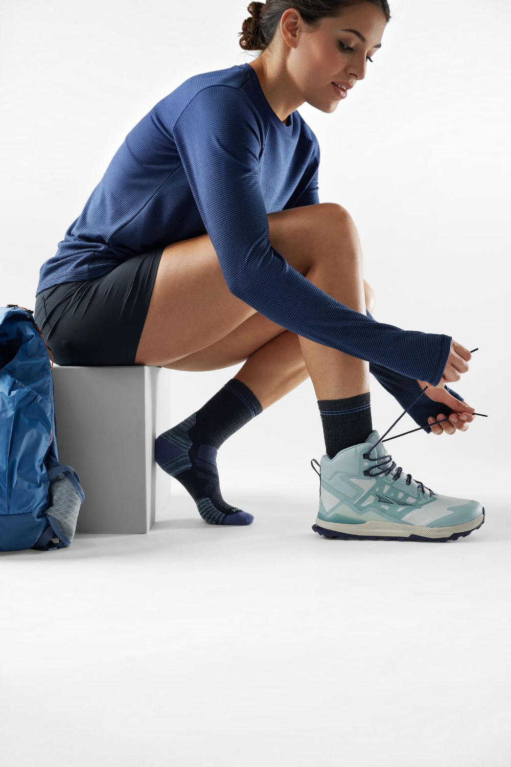 A woman in a navy blue long-sleeve athletic top, black shorts, charcoal socks with purple accents, and a light blue and gray trail running shoe sits on a gray block, tying the laces of her shoe. A blue backpack rests on the floor beside her against a clean white studio background.