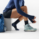 A woman in a navy blue long-sleeve athletic top, black shorts, charcoal socks with purple accents, and a light blue and gray trail running shoe sits on a gray block, tying the laces of her shoe. A blue backpack rests on the floor beside her against a clean white studio background.