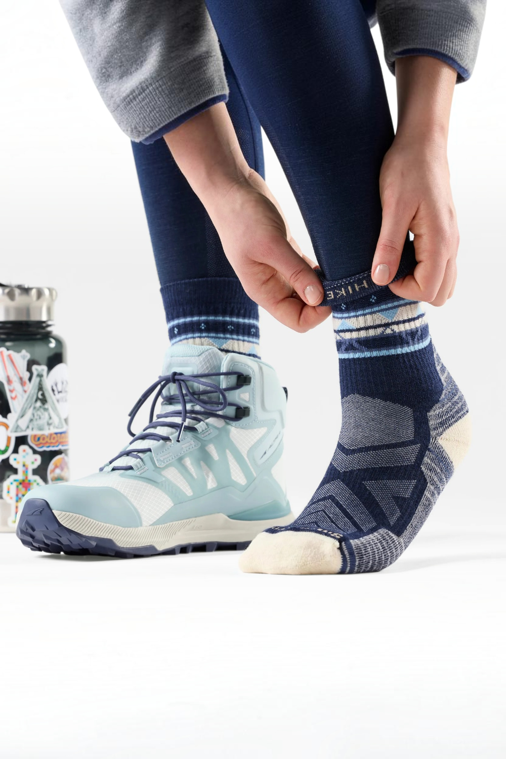 Person wearing navy leggings and a grey sweatshirt putting on a navy and cream mixed pattern sock. The other foot already has a hiking boot on.
