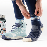Person wearing navy leggings and a grey sweatshirt putting on a navy and cream mixed pattern sock. The other foot already has a hiking boot on.