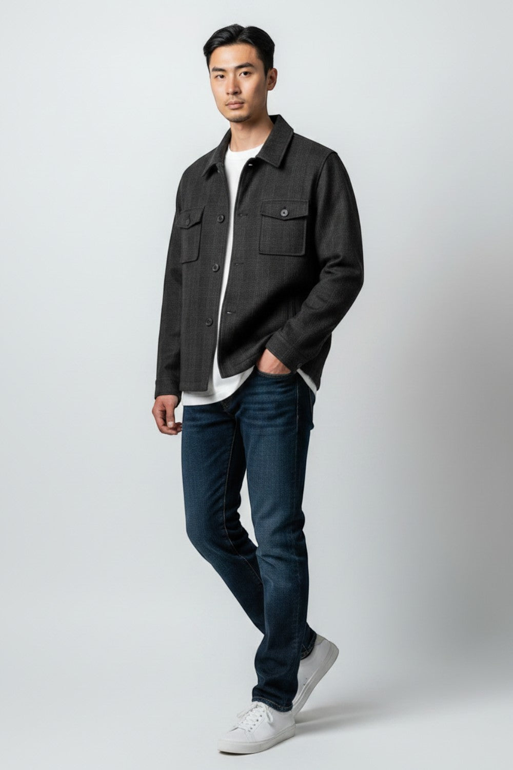 A man modeling a dark charcoal gray overshirt jacket over a white t-shirt, paired with dark blue jeans and white sneakers, standing against a light gray studio background.