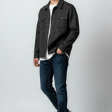A man modeling a dark charcoal gray overshirt jacket over a white t-shirt, paired with dark blue jeans and white sneakers, standing against a light gray studio background.