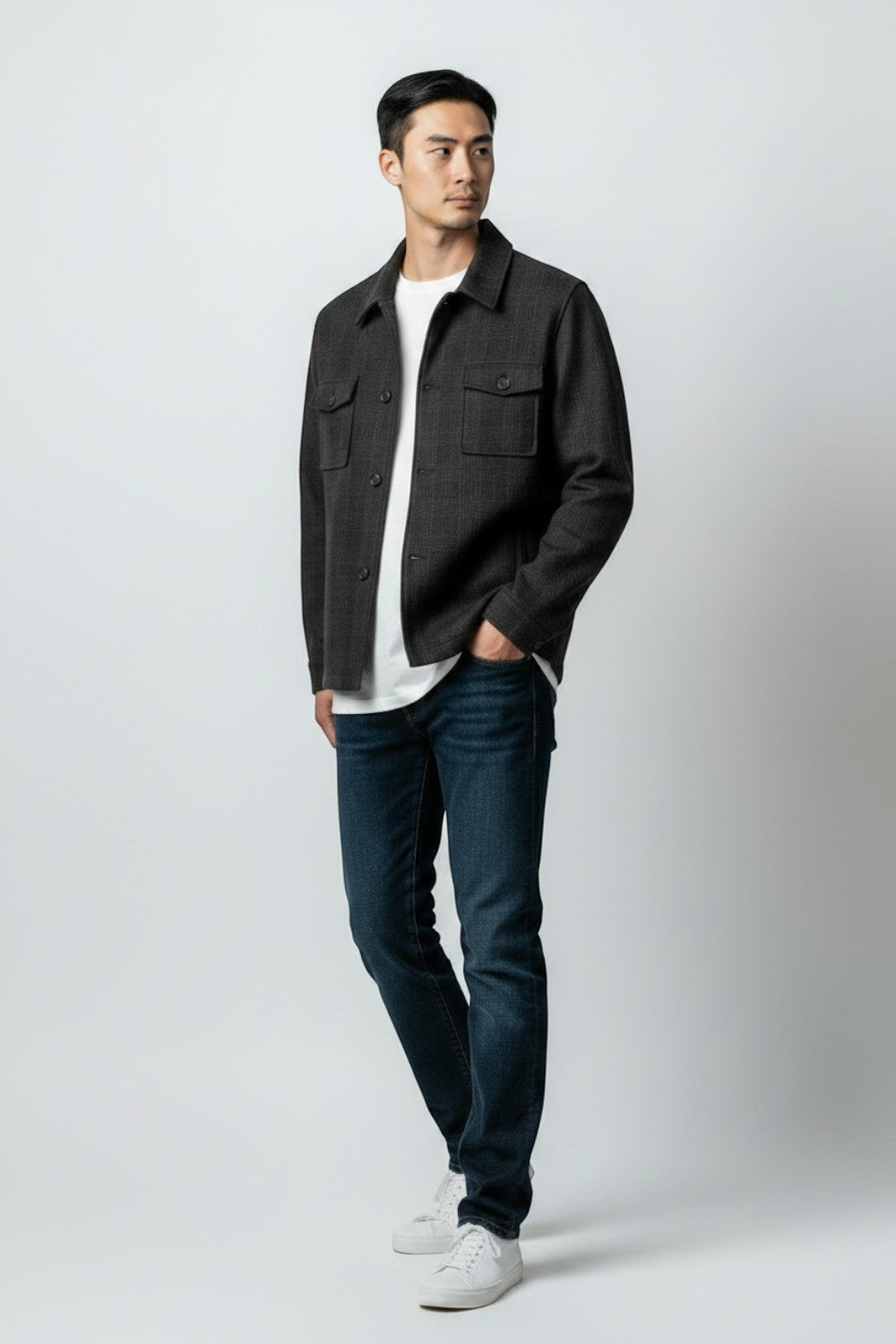 A man modeling a dark charcoal gray textured overshirt jacket over a white t-shirt, paired with dark blue jeans and white sneakers, standing against a light gray studio background.