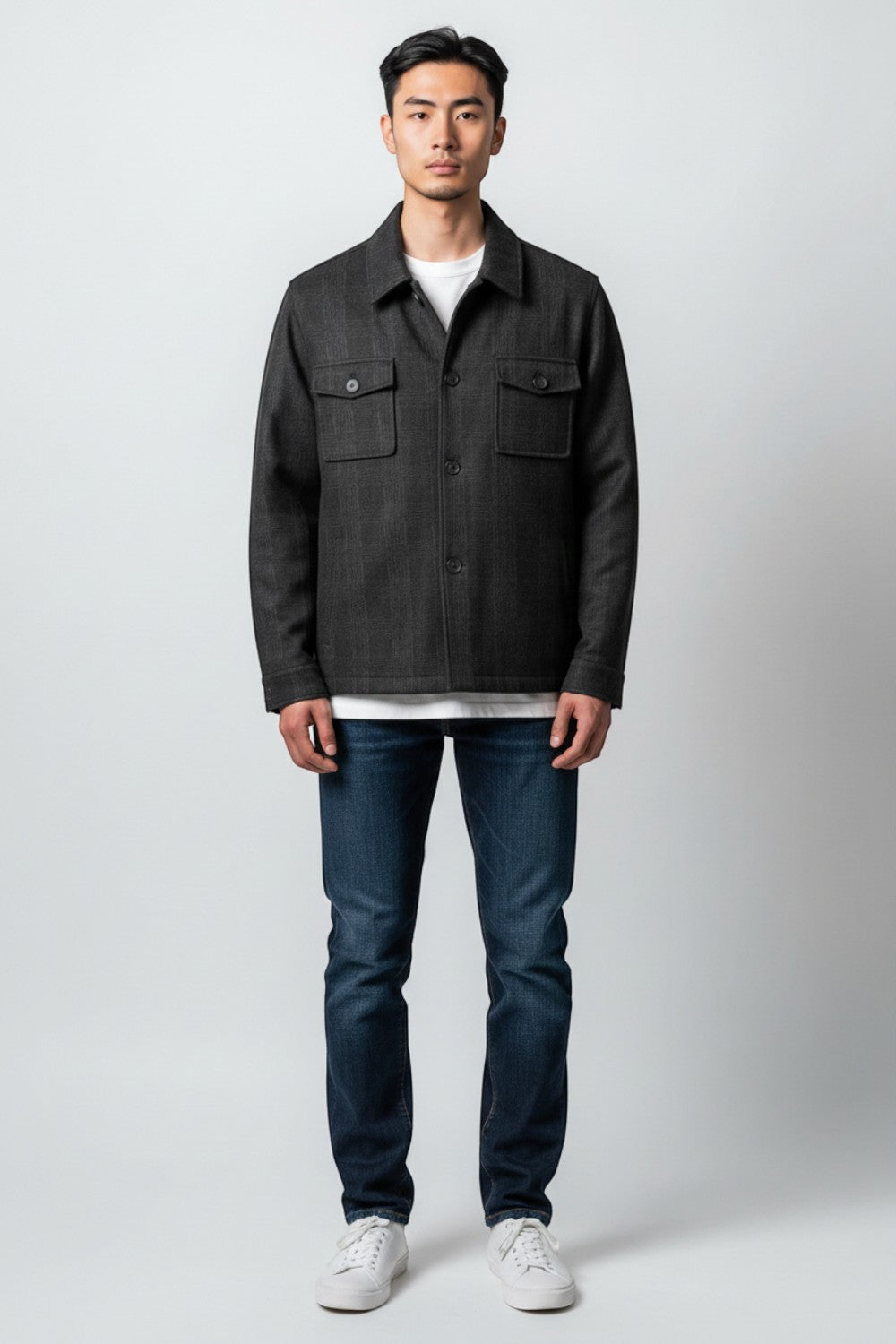 A man modeling a dark charcoal gray textured overshirt jacket over a white t-shirt, paired with dark blue jeans and white sneakers, standing against a light gray studio background.