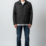 A man modeling a dark charcoal gray textured overshirt jacket over a white t-shirt, paired with dark blue jeans and white sneakers, standing against a light gray studio background.
