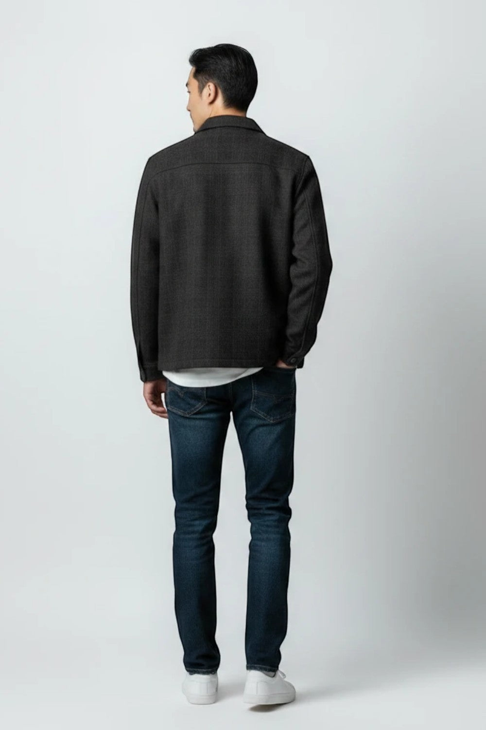 A man shown from the back wearing a dark charcoal gray textured jacket, dark blue jeans, and white sneakers, standing against a light gray studio background.