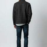 A man shown from the back wearing a dark charcoal gray textured jacket, dark blue jeans, and white sneakers, standing against a light gray studio background.