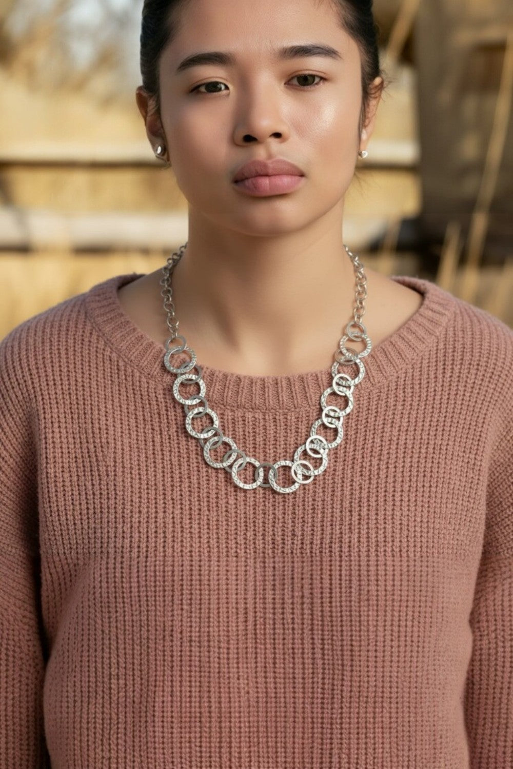 Woman wearing a pink sweater and silver chain necklace with a blurred outdoor background