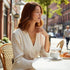 Woman sitting at an outdoor cafe table with a cup of coffee and croissant, surrounded by greenery and buildings.