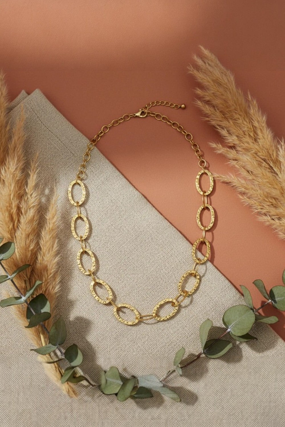 Gold chain necklace on a neutral fabric background with pampas grass and eucalyptus leaves.