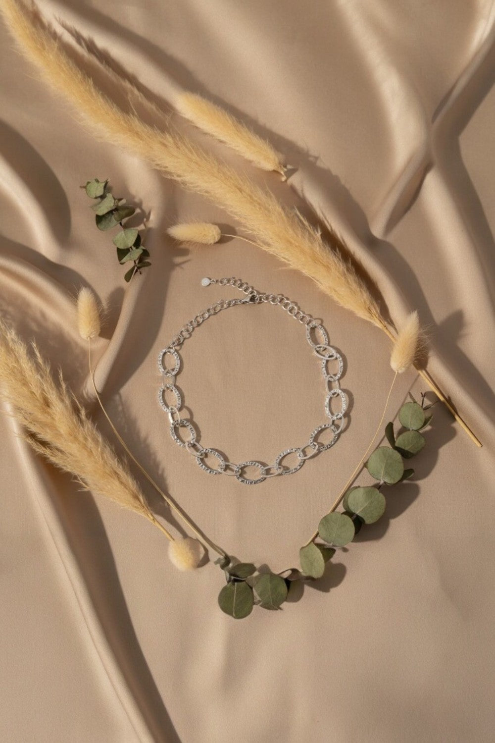 Silver chain necklace on a beige fabric background with dried grass and green leaves.