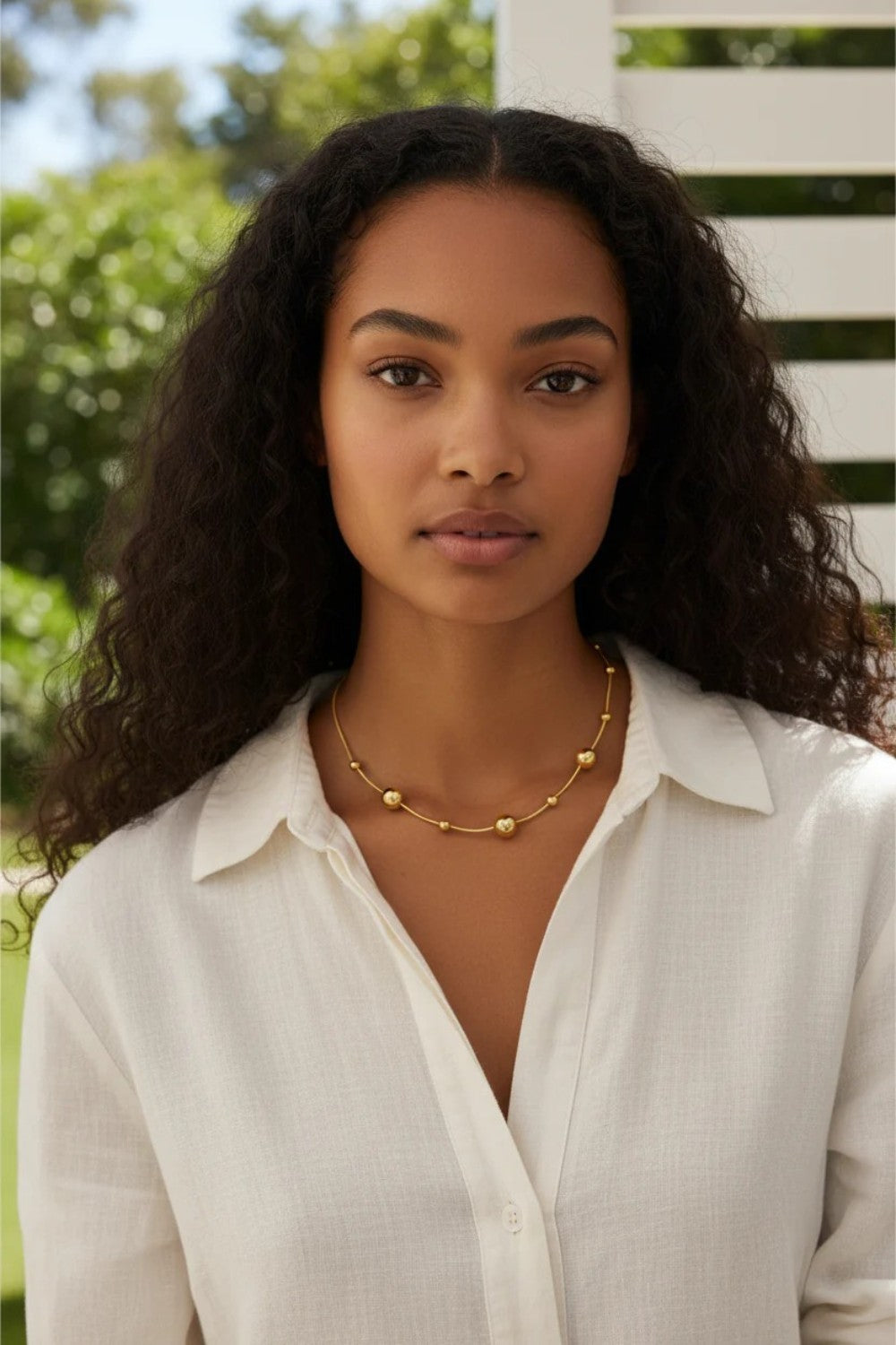 Woman wearing a gold necklace outdoors with greenery in the background