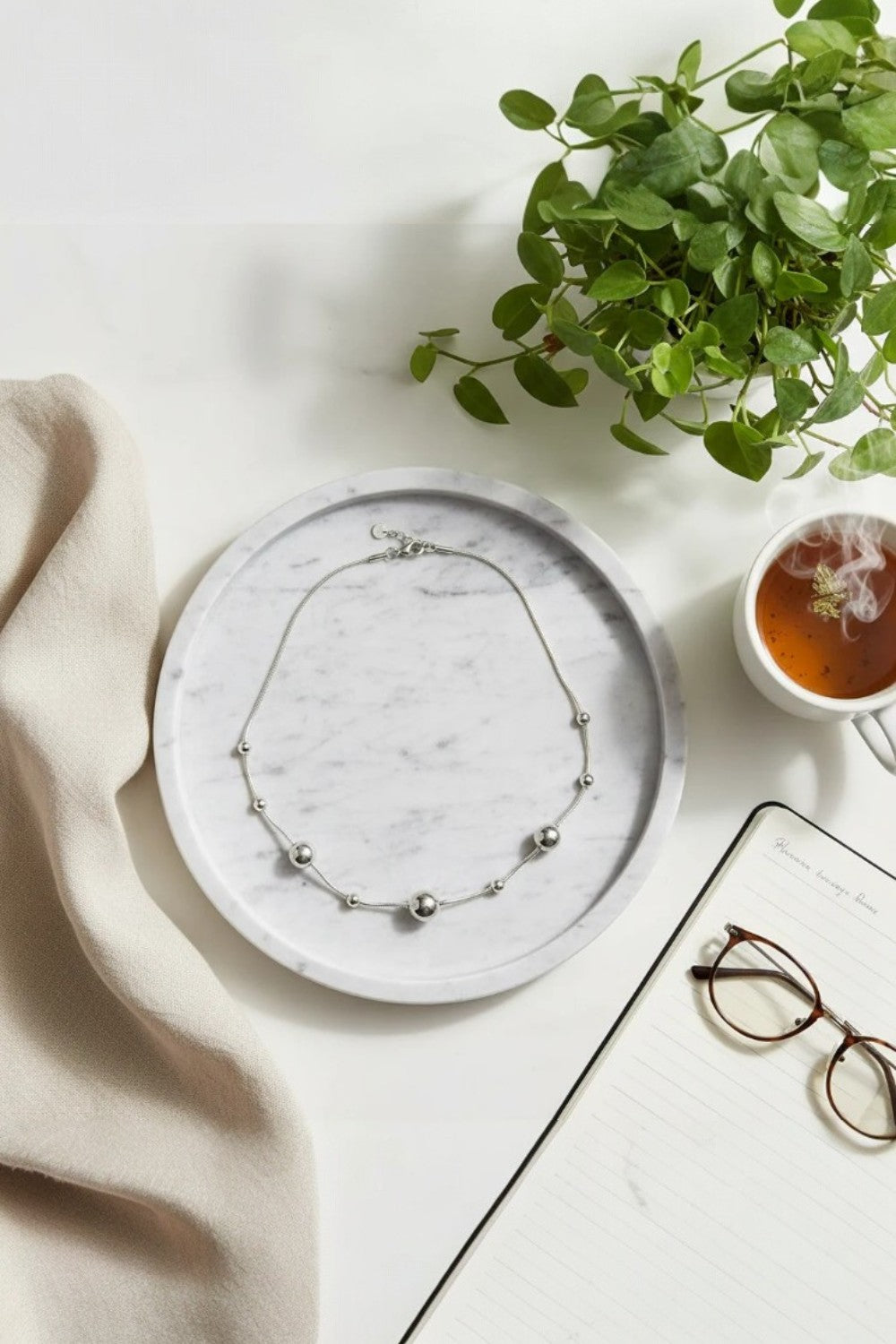 Necklace on a marble plate with a plant, tea cup, and glasses on a light surface