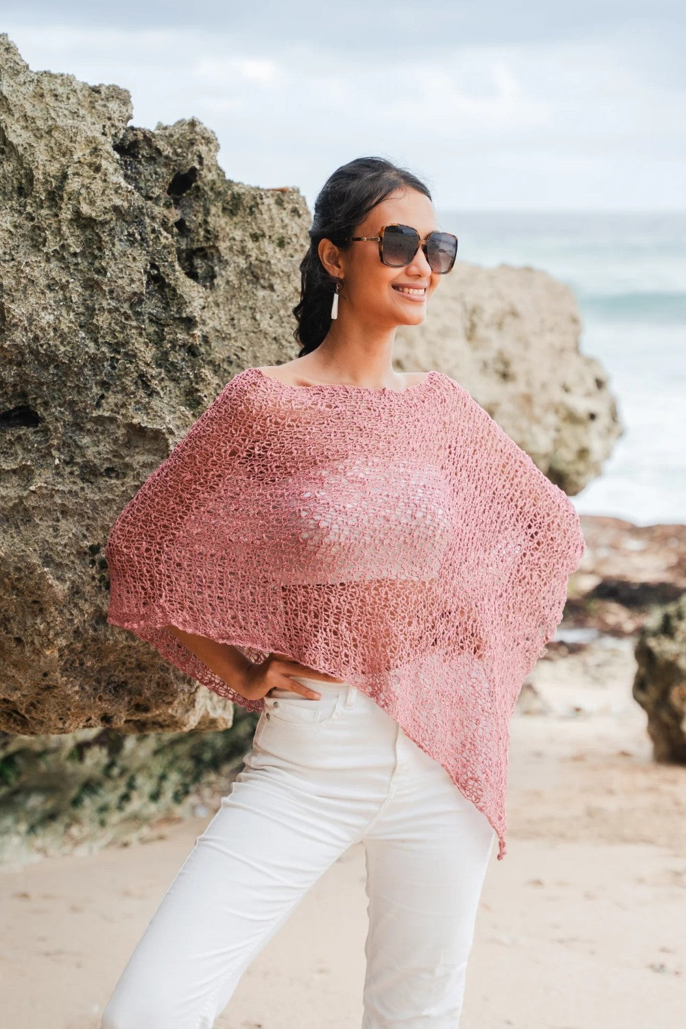 Woman wearing a pink knitted poncho and sunglasses on a beach.