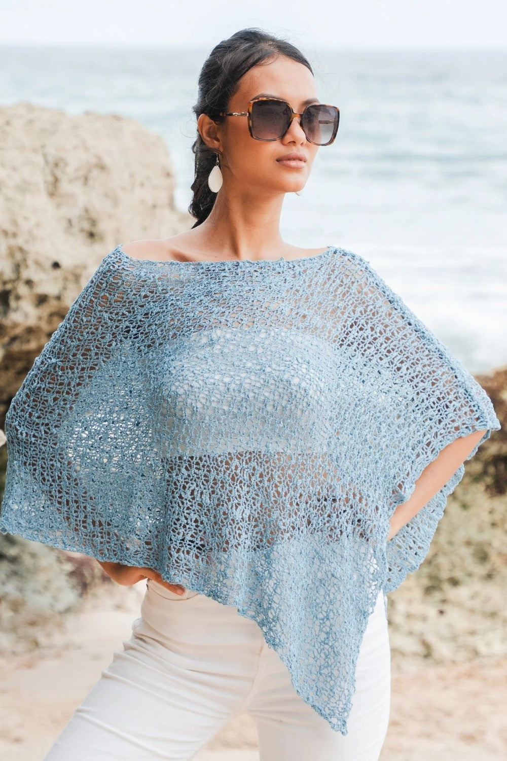Woman wearing a light blue crochet poncho with white pants in an outdoor setting