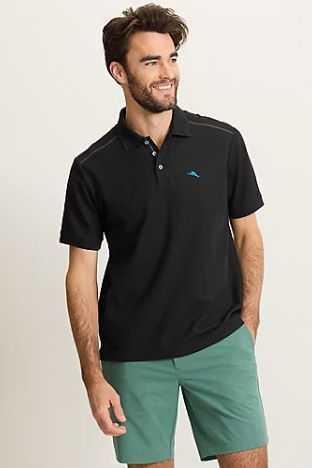 Man wearing a black polo shirt with a small logo and green shorts on a beige background