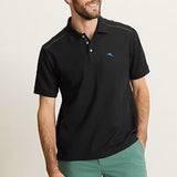 Man wearing a black polo shirt with a small logo and green shorts on a beige background