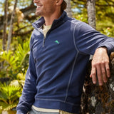 Man wearing a blue pullover with a logo, sunglasses, and a cap in a natural setting.