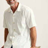 Man wearing a white short-sleeve shirt sitting on a wooden stool against a plain background