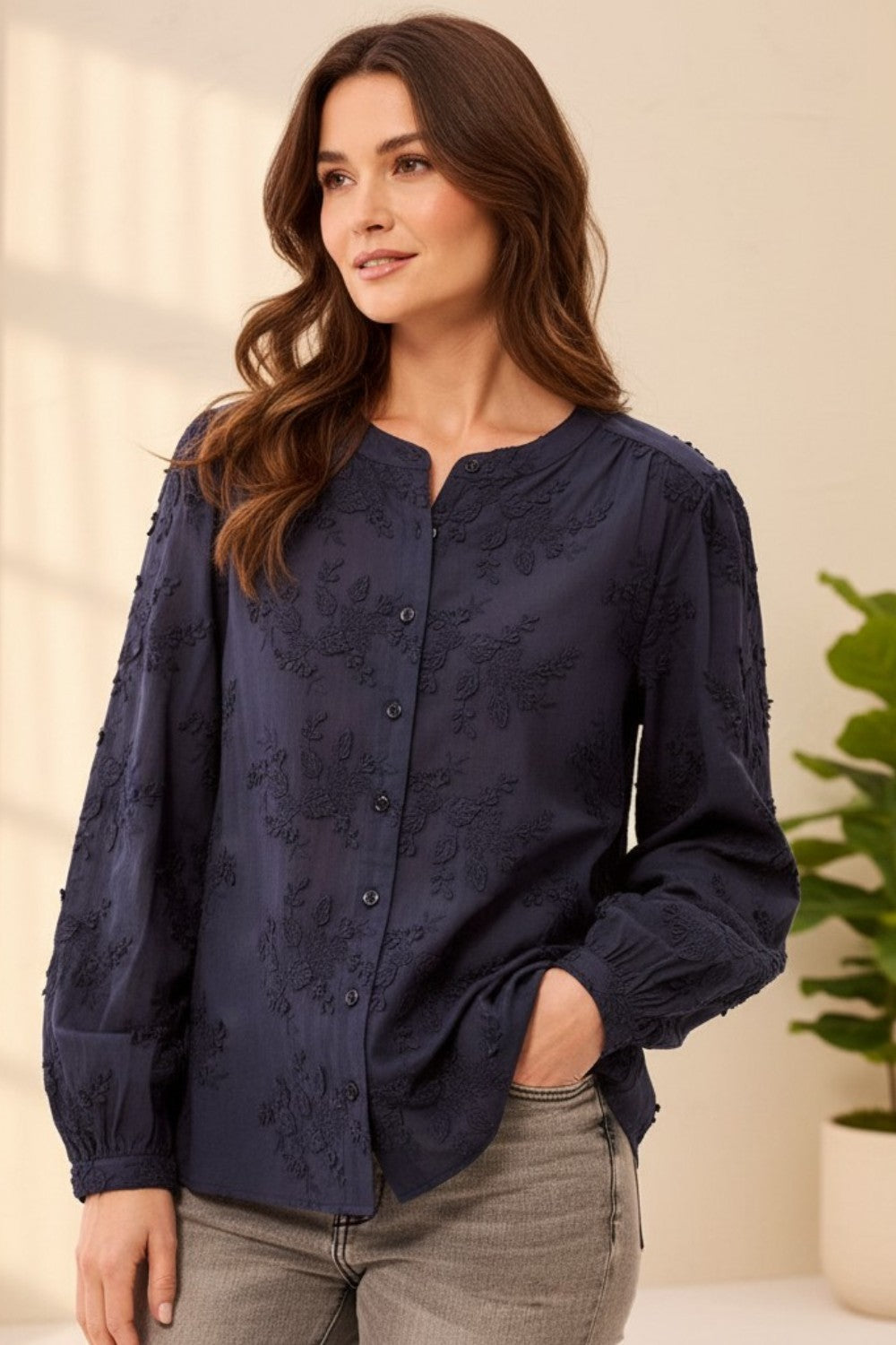 A woman wearing a navy blue long-sleeve blouse with tonal floral embroidery and a button-down front, paired with gray jeans, in a brightly lit indoor setting.