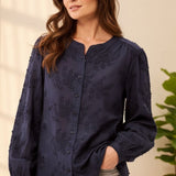 A woman wearing a navy blue long-sleeve blouse with tonal floral embroidery and a button-down front, paired with gray jeans, in a brightly lit indoor setting.