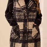 Woman wearing a black and tan plaid vest with a button front and functional pockets over a black dress against a light backdrop.