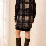 Woman wearing a black and tan plaid vest over a black dress with tall brown boots against a light backdrop.