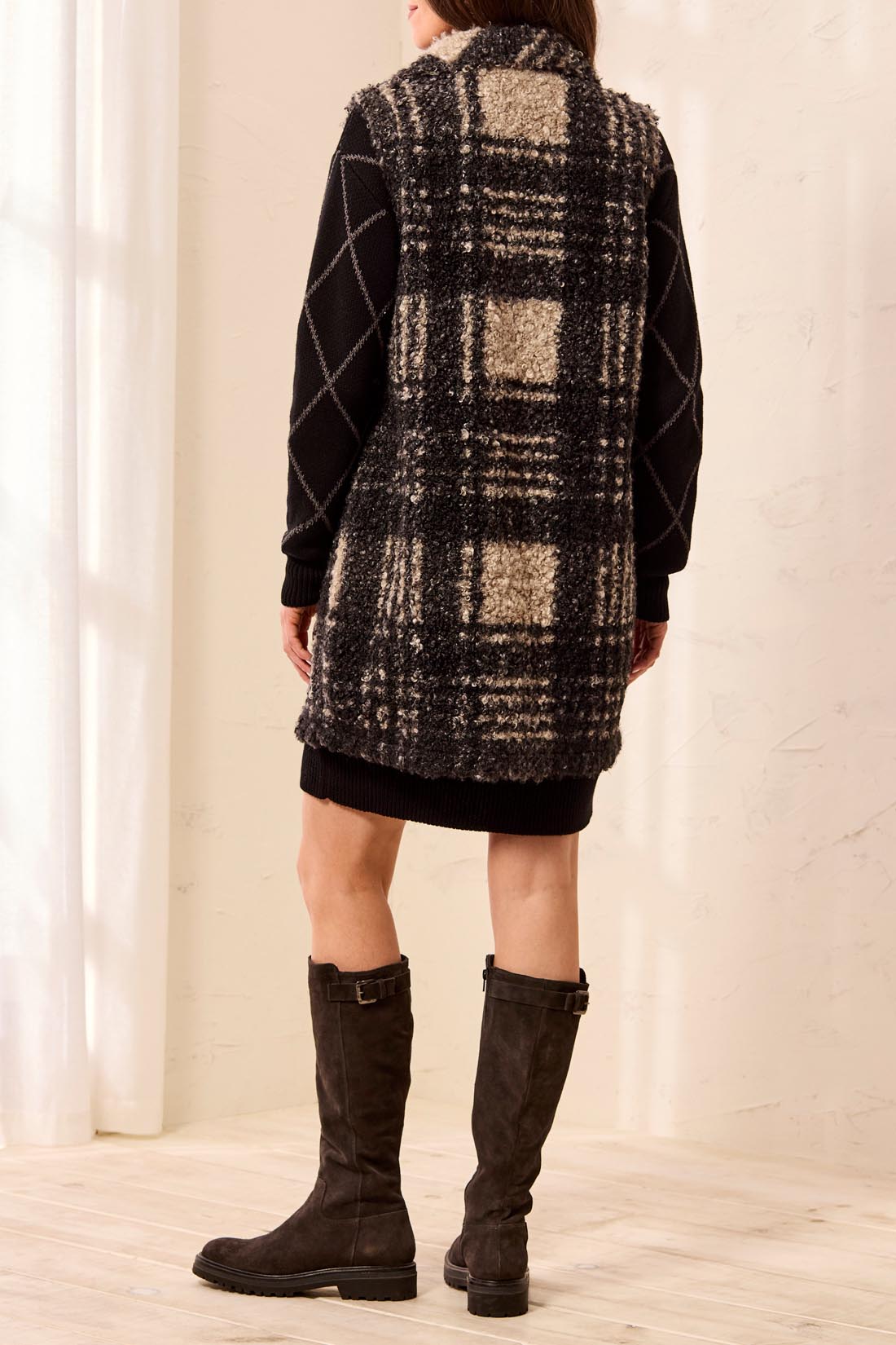 Woman wearing a black and tan plaid vest over a black dress with tall brown boots against a light backdrop.