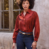 A woman with an afro hairstyle models a rust-colored long-sleeve shirt with a wide fringed yoke, paired with dark blue jeans and a brown belt, standing against a textured stucco wall and rustic window.